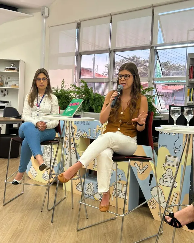 Bianca Salles sentada num banco alto, ao microfone, num espaço do Senac, com um exemplar do livro exposto num cavalete ao lado.