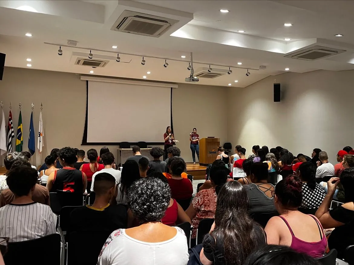 Auditório visto do fundo da sala, com a plateia sentada de costas para a câmera e Bianca Salles ao microfone no palco, ao lado de um púlpito do Senac.